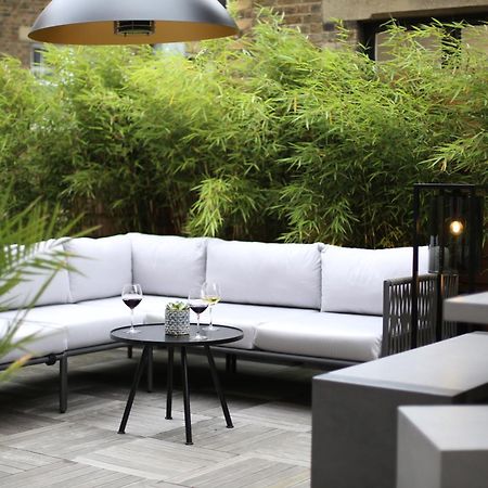 Middle Eight - Covent Garden - Preferred Hotels And Resorts Лондон Екстериор снимка The photo shows an outdoor seating area featuring a modern gray sectional sofa with plush cushions. In front of the sofa is a small, round black table with two glasses of wine on it, accompanied by a decorative plant or vase. Surrounding the area are