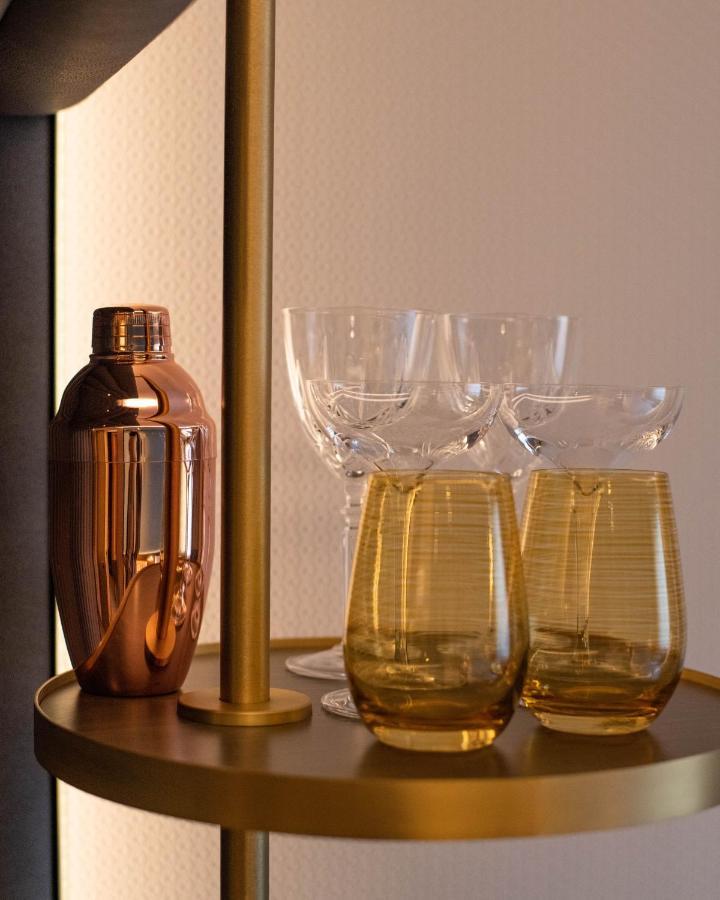 Middle Eight - Covent Garden - Preferred Hotels And Resorts Лондон Екстериор снимка The photo shows a stylish bar setup featuring a shiny copper cocktail shaker positioned next to several elegant glassware pieces. There are four tall, clear stemmed glasses and two round, amber-colored glasses with a textured pattern. The background 
