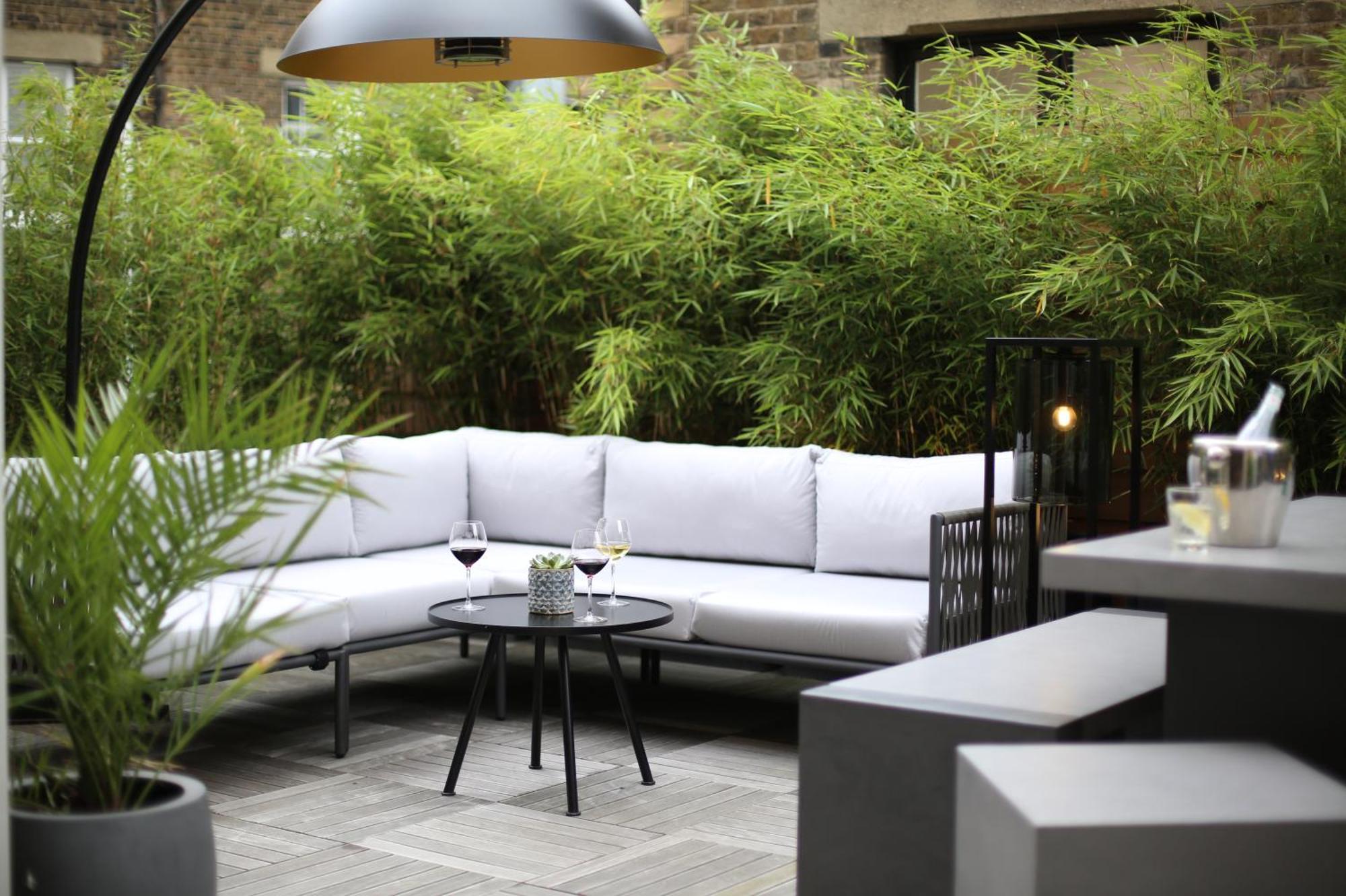 Middle Eight - Covent Garden - Preferred Hotels And Resorts Лондон Екстериор снимка The photo shows an outdoor seating area featuring a modern gray sectional sofa with plush cushions. In front of the sofa is a small, round black table with two glasses of wine on it, accompanied by a decorative plant or vase. Surrounding the area are